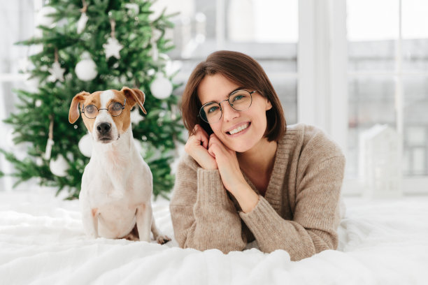 美丽的黑发成年女子戴着大圆眼镜，斜着头，穿着温暖的冬衣，她的眼镜宠物在床边摆姿势，表达快乐的情绪，等待圣诞节，享受舒适图片下载