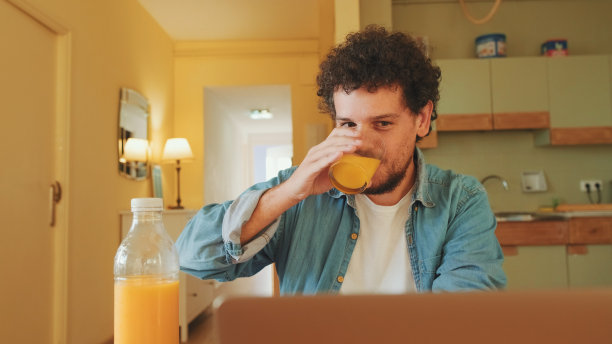 一个穿着牛仔衬衫的人坐在厨房里一边喝酒一边用笔记本电脑工作图片下载