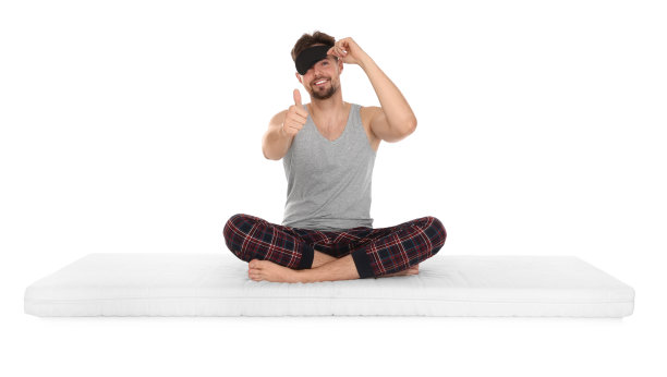 戴着睡眠面罩的微笑男子坐在柔软的床垫上，在白色的背景下竖起大拇指图片下载