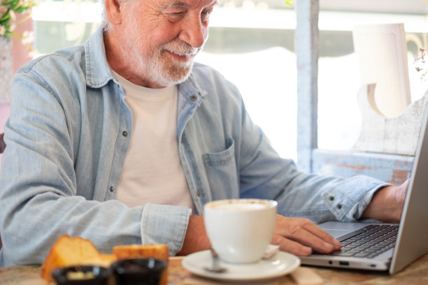 一个面带微笑的老人在咖啡店里一边用笔记本电脑工作，一边喝着卡布奇诺和甜蛋糕的早餐。老年人享受自由的生活方式和愉快的放松时刻图片下载