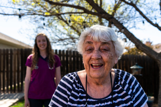 老妇人对着镜头微笑，她的女护士骄傲地站在她身后，也微笑着。这是一个温暖人心的时刻，他们之间充满了感情和信任。图片下载