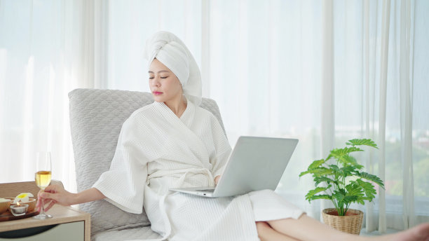 周末在豪华卧室里休息时，穿着浴袍和毛巾的亚洲女性在家里的沙发床上使用笔记本电脑图片下载