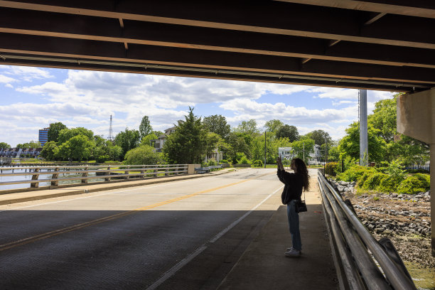 周末在汉普顿散步，在桥下拍照。年轻的棕色头发的白人女人拿着智能手机在小镇的桥下散步图片下载