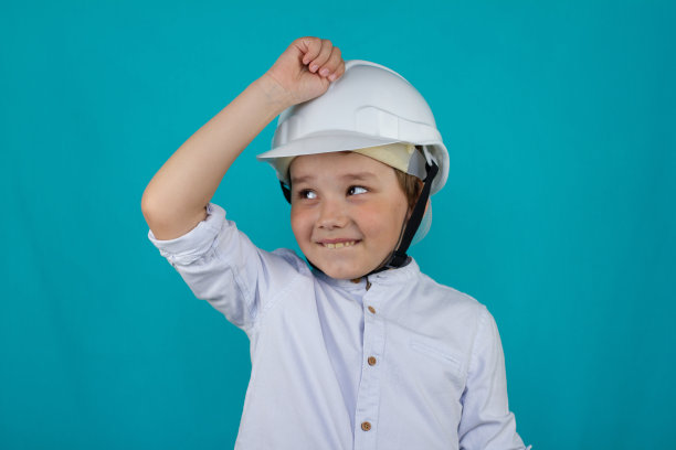 一个可爱的孩子戴着建筑头盔在开玩笑。孩子用拳头敲打着头上的施工头盔。男孩滑稽地用拳头敲了一下头盔，笑了。图片下载