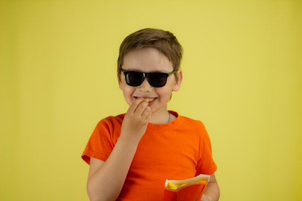 孩子微笑着吃着黄色背景的炸薯条。这个男孩手里拿着他最喜欢的食物炸薯条。男孩咬下一片薯条，看着镜头笑了。图片下载