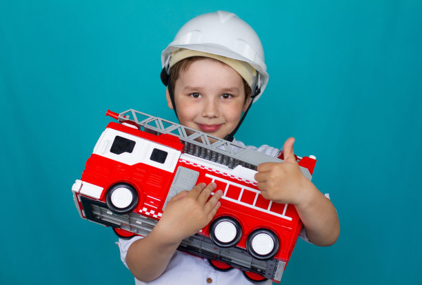 一个戴安全帽的男孩手里拿着一辆玩具消防车，微笑着竖起大拇指。孤立在蓝色背景上。这个男孩梦想成为一名消防员。概念儿童与消防车。图片下载