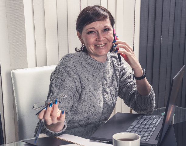快乐的女商人在办公室里对着笔记本电脑讲手机。成功的事业，人们微笑着工作图片下载