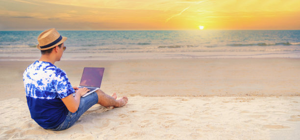 年轻的亚洲人在度假期间坐在美丽的海滩上，用笔记本电脑在户外工作。夏天。假期。度假和快乐的人的概念。图片下载