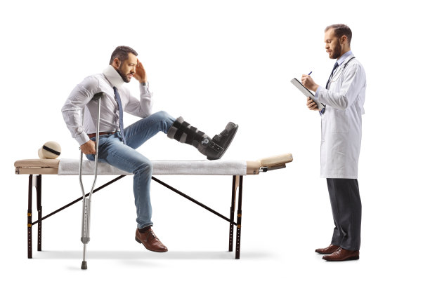 一个受伤的男人，穿着矫形靴，戴着颈圈，坐在治疗台上，医生正在写文件图片下载