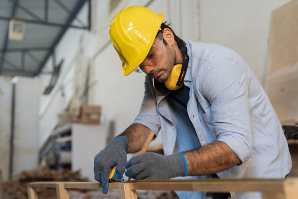 男木匠在木工车间测量木材的尺寸。在家具车间工作的男性细木工，穿着安全工作服，戴着手套和安全帽图片下载