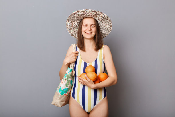 开朗、快乐、积极的女人穿着泳装，孤立在灰色的背景下，站在水果旁边，看着相机享受她的假期。图片下载