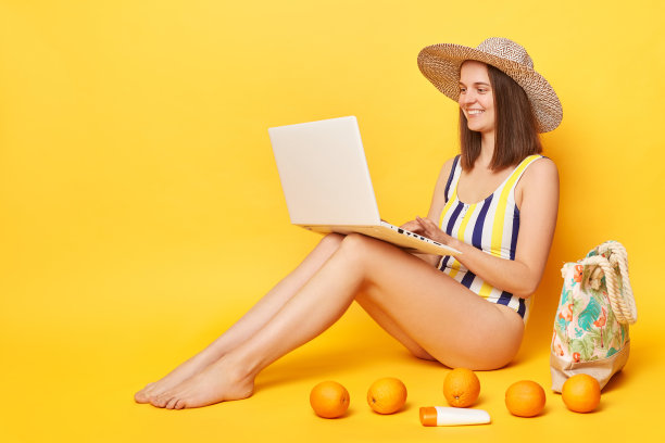 努力工作的有魅力的女人穿着一件泳衣，在黄色的背景下摆出孤立的姿势，在度假胜地工作，坐在笔记本电脑前享受她的远程工作。图片下载