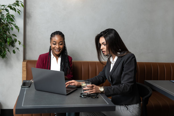 一对一的会议。两个年轻的女商人坐在咖啡馆的桌子旁。女孩在笔记本电脑屏幕上展示同事信息。女孩用智能手机，写博客。团队合作，商务会议。自由职业者的工作。图片下载