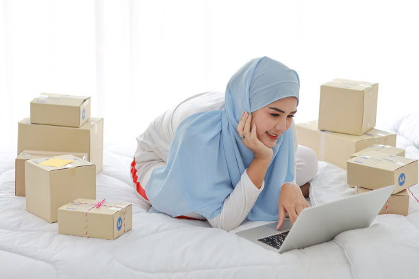 美丽和年轻的亚洲妇女穿着穆斯林睡衣，有吸引力的样子，躺在床上的电脑和在线包裹盒交付。创业中小企业自由职业女孩用手机工作图片下载