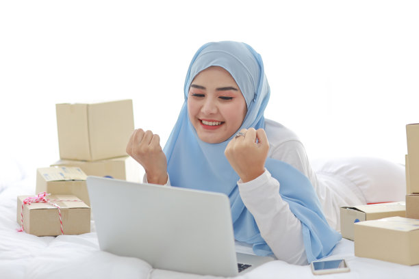 年轻美丽的亚洲女子穿着穆斯林睡衣，外表迷人，躺在床上，电脑，手机和在线包裹箱送货。戴头巾的聪明女孩收到好消息和惊喜。图片下载