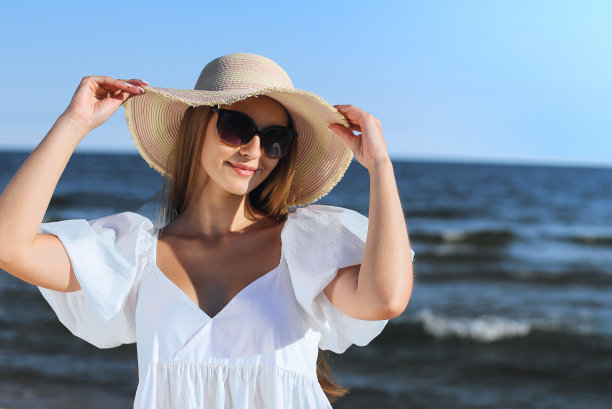快乐的金发女郎戴着太阳镜和帽子在海边摆姿势。晚上的太阳图片下载