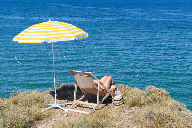 女人戴着草帽，坐在日光浴床上晒太阳，躺椅下的黄伞在山顶，悬崖与蓝色的大海，海景。图片下载