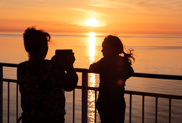 一家人，一个年轻的女人和一个孩子站在船的甲板上，在夕阳的映衬下拍照。小女孩和女人的剪影。图片下载