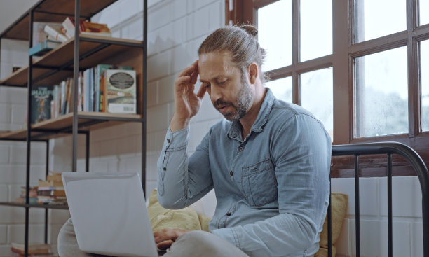 忧心忡忡的年轻人坐在卧室的床上使用笔记本电脑。图片下载