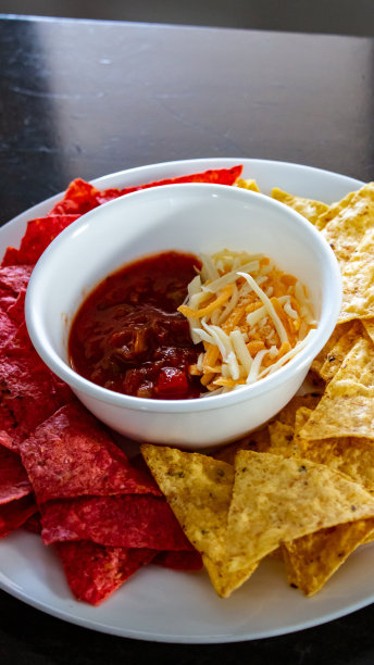 白色的盘子和碗放在棕色的桌子上，有红色和黄色的玉米饼片、莎莎酱和奶酪图片下载