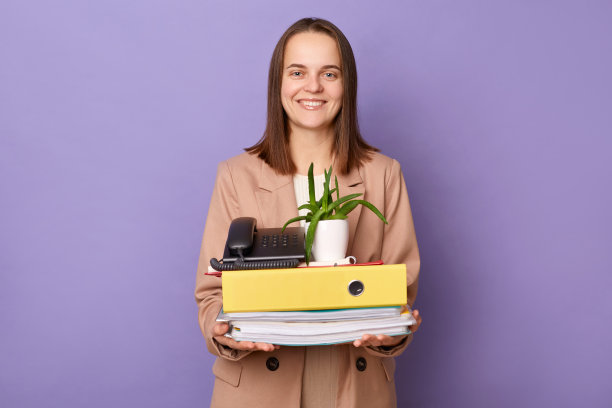 微笑开朗的女办公室职员，拿着装着她的东西的纸板箱，找到了新工作，看着相机，幸福地孤立在紫色的背景上。图片下载