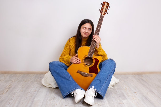一个穿着牛仔裤和毛衣的年轻女孩坐在地板上，手里拿着一把原声吉他。图片下载