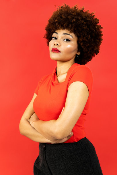 一个深色皮肤的女孩，留着非洲式发型，在红色背景上摆姿势的垂直照片。这个女孩双臂交叉摆姿势，一副悲伤的表情。悲伤，悲伤或抑郁的概念。图片下载