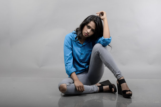 印度女年轻模特在休闲服装对灰色背景-股票照片。黑色长发模特穿着蓝色衬衫，灰色牛仔裤和黑色高跟鞋。图片下载