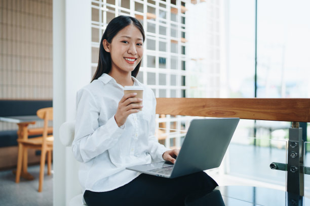 一位年轻的亚洲个体户妇女在咖啡店拿着咖啡杯，一边使用电脑一边露出微笑和快乐。自雇、自由职业、放松。图片下载