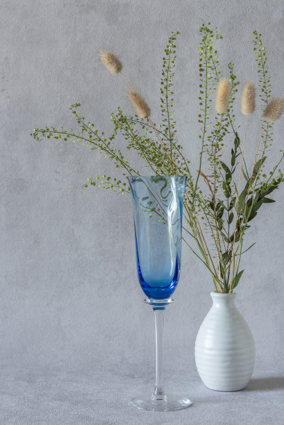 蓝色的葡萄酒杯和花瓶与干植物在灰色的背景。副本的空间。图片下载