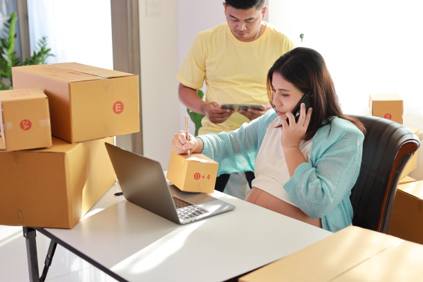 亚洲夫妇进行箱子包装工作，年轻孕妇坐着使用平板电脑和智能手机记录客户订单，丈夫一同工作图片下载