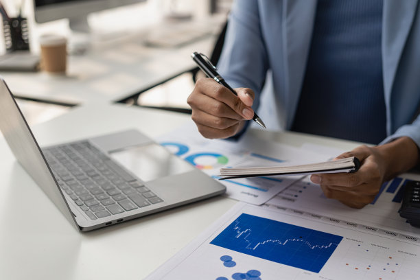 在办公室里，坐在木桌前用笔记本电脑工作的年轻女性是一名商人，她分析文件和图表，用笔记本电脑和计算器工作。远程图片图片下载