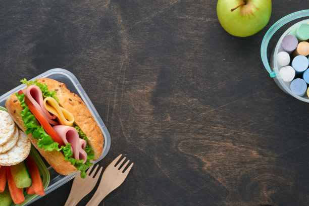 学校午餐的健康食品概念。健康面包，肉三明治，奶酪，苹果，新鲜黄瓜，胡萝卜，坚果在容器里，水和酸奶在黑暗的背景。回到学校的概念。图片下载