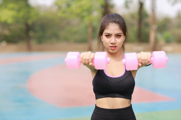 运动年轻美丽的亚洲女子，穿着运动服站在户外举哑铃做晨练。活跃的女孩在绿树背景下锻炼二头肌。体育的概念图片下载