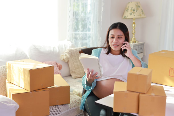 迷人快乐的亚洲孕妇坐着使用移动智能手机，用平板电脑为客户写订单。准妈妈在怀孕期间为婴儿的出生做准备和工作。图片下载