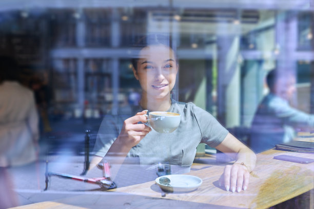 年轻女性在咖啡馆里拿着一杯咖啡，透过窗户玻璃往外看图片下载