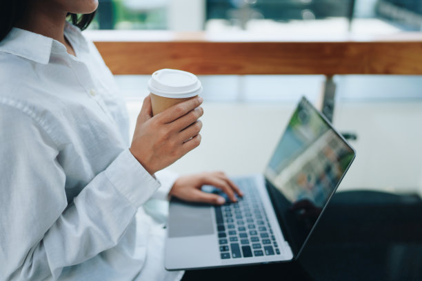 一位年轻的亚洲个体户妇女在咖啡店拿着咖啡杯，一边使用电脑一边露出微笑和快乐。自雇、自由职业、放松。图片下载