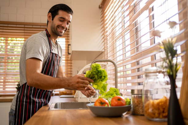 快乐的年轻西班牙裔男人穿着灰色衬衫和围裙在厨房里洗西红柿图片下载