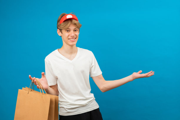 年轻的卖家穿着休闲的白色t恤，带着纸袋，孤立在蓝色背景的工作室肖像上。人们的生活理念。模拟复制空间。十几岁的孩子拿着装着买的东西的包图片下载