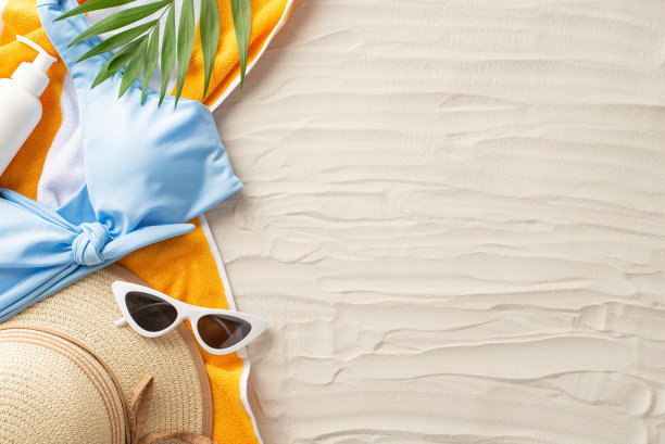 拥抱夏天的炎热。俯看日光浴必备物品:太阳镜、帽子、蓝色女式泳衣、防晒霜、黄色毛巾、棕榈叶。沙滩设置，为文本或广告留出空白空间图片下载