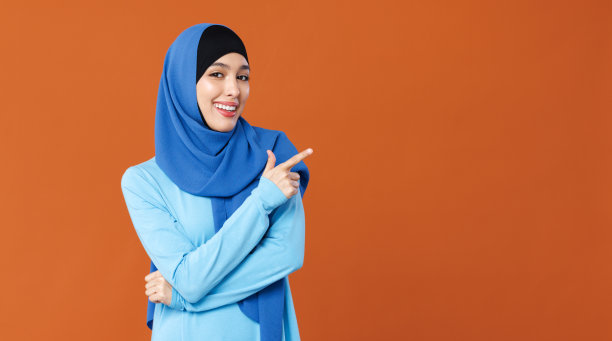 年轻貌美的穆斯林妇女面带微笑，头戴传统宗教头巾，在橙色背景下指着一边图片下载