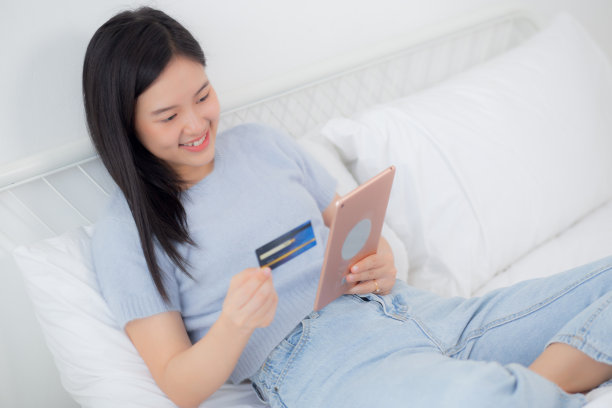 年轻的亚洲女性坐在床上用数码平板电脑在家里的卧室用信用卡网上购物，女性支付用交易金融，购买和支付，商业理念。图片下载