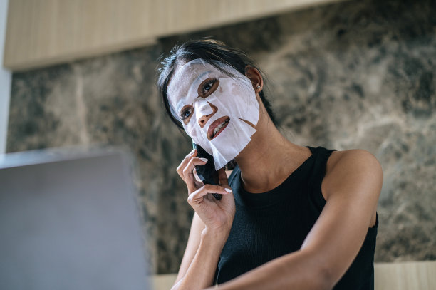 年轻的亚洲女性戴着面膜坐在家里的床上，一边用笔记本电脑工作图片下载