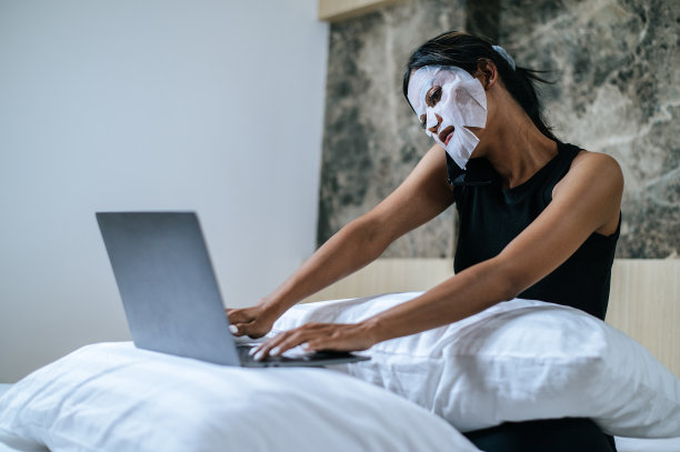 年轻的亚洲女性戴着面膜坐在家里的床上，一边用笔记本电脑工作图片下载