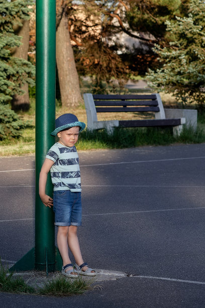 小男孩在公园迷路了。一个生气的孩子站在操场上的一根绿杆子旁边。一个孤独的孩子在公园里哭泣图片下载