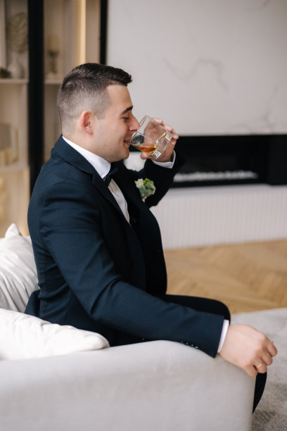英俊的新郎穿着深蓝色西装，端着一杯威士忌坐在椅子上。新郎的早晨。结婚的那一天图片下载