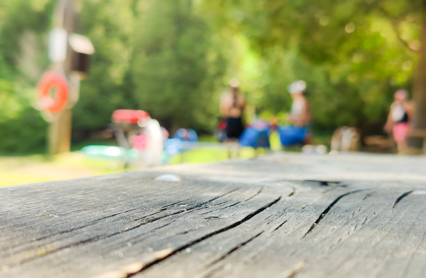 在一个阳光明媚的户外公园里，前景以木凳为特色，而背景则是一个模糊的绿色景观，人们在草地上野餐和嬉戏。图片下载