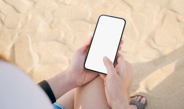 手持智能手机在沙滩上，手机模型的空白屏幕，拿你的屏幕投放广告。暑假概念。图片下载