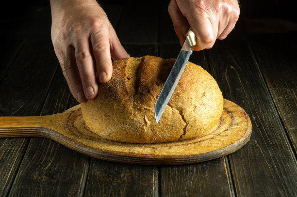 小麦面包放在刚从烤箱里拿出来的砧板上。厨师手里拿着刀切新鲜的全麦面包图片下载
