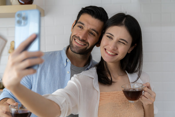 疲惫的亚洲管家在休息时自拍，用手机，年轻夫妇对着相机拥抱微笑，坐在地板上，周围是清洁工具和用品，厨房内部。图片下载